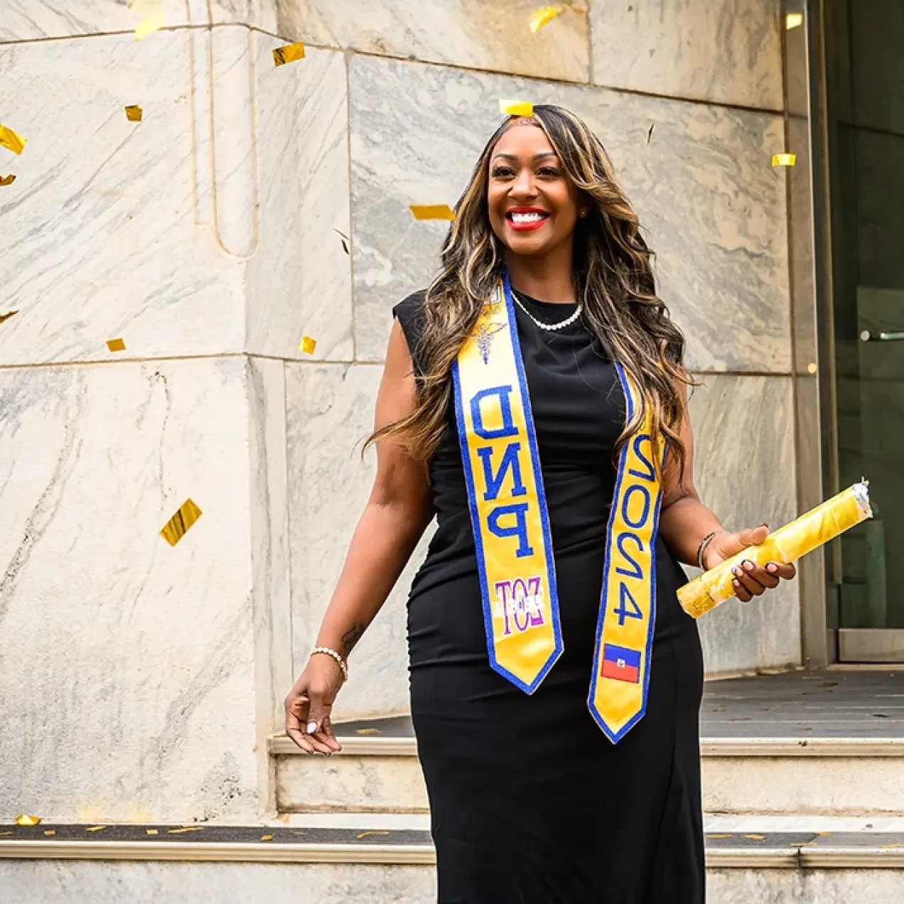 Graduating student celebrating with confetti, wearing a black dress and DNP 2024 sash, holding a diploma outside a building.